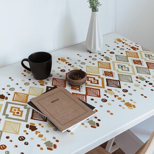 Op de afbeelding: Een wit bureau met een geometrisch patroon dat op het oppervlak is gedrukt. Het patroon bevat bruine, oranje en groene vierkanten en cirkels. Een bruin notitieboek met het woord "NOTES" op de omslag ligt op het bureau, samen met een zwarte koffiemok en een kleine kom met paperclips.