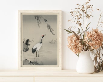Op de afbeelding: Een ingelijste zwart-witprint van een kraanvogel die in een beek staat met een wilg erboven. De print is op een lichtgrijze achtergrond en heeft een witte lijst.
