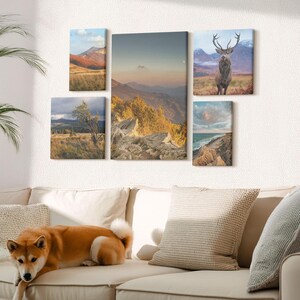 Peut inclure: Un ensemble d'art mural sur toile représentant des paysages. Les images montrent des montagnes, une scène côtière et un cerf. La palette de couleurs comprend des tons de terre, du bleu et du vert. L'œuvre est affichée sur un mur blanc au-dessus d'un canapé beige.