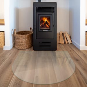 May include: A clear, semi-circular glass hearth plate positioned on a wooden floor in front of a black metal wood-burning stove. The stove features a glass door revealing burning logs with orange flames, and a woven basket sits to the left.