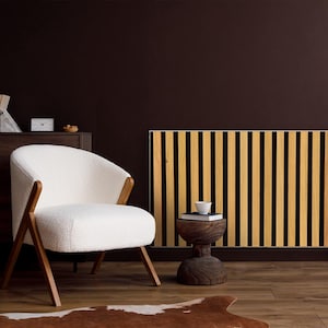 Op de afbeelding: Een witte fauteuil met houten poten staat voor een houten radiator met zwarte en bruine verticale strepen. Een kleine houten kruk met een kopje en een boek staat voor de radiator.