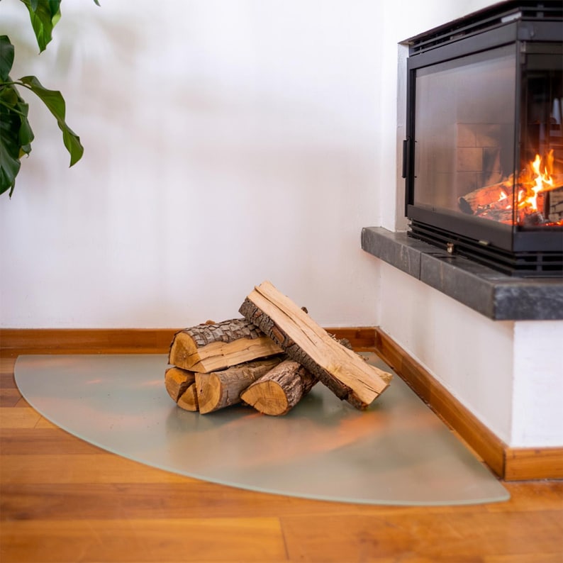 May include: A semi-circular, frosted glass hearth pad placed on a wooden floor in front of a fireplace. A small stack of split, light brown firewood logs with textured bark rests directly on the centre of the glass floor protector.