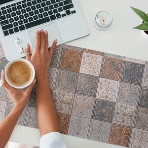 Könnte beinhalten: Nahaufnahme eines Schreibtisches mit einer gemusterten Schreibtischunterlage, die ein sich wiederholendes Muster aus verzierten Fliesen in Braun-, Grau- und Weißtönen zeigt. Ein Laptop, eine Tasse Kaffee und eine kleine Topfpflanze stehen auf dem Schreibtisch.