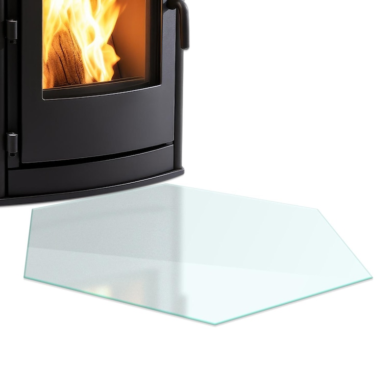 May include: A clear, hexagonal glass floor plate positioned in front of a black metal wood-burning stove. The stove features a glass door revealing bright orange flames and burning logs inside, with a curved front design and a black handle.