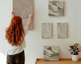 Puede incluir: Una colección de obras de arte murales texturizadas en tonos neutros. El arte presenta diseños abstractos, incluyendo patrones topográficos y formas fluidas en forma de ola. Las piezas se exhiben en una pared blanca, con un mueble de madera y una planta en maceta.