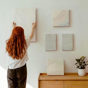 May include: A person hanging a large, rectangular, off-white canvas on a white wall. Several smaller square and rectangular canvases with abstract designs are already mounted. A wooden cabinet with a potted plant sits below the artwork, creating a gallery-like display.