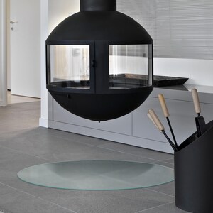 May include: A matte black, spherical, suspended fireplace featuring a glass-enclosed firebox and a central chimney pipe. Below the fireplace sits a circular glass floor plate. A black metal tool holder with light wood handles is positioned nearby.