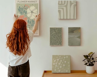 Può includere: Una donna appende un poster "FLOWER MARKET" con arte floreale. Quattro opere d'arte su tela quadrate e rettangolari con disegni astratti e una piccola pianta in vaso sono esposte sulla parete e sul mobile in legno.