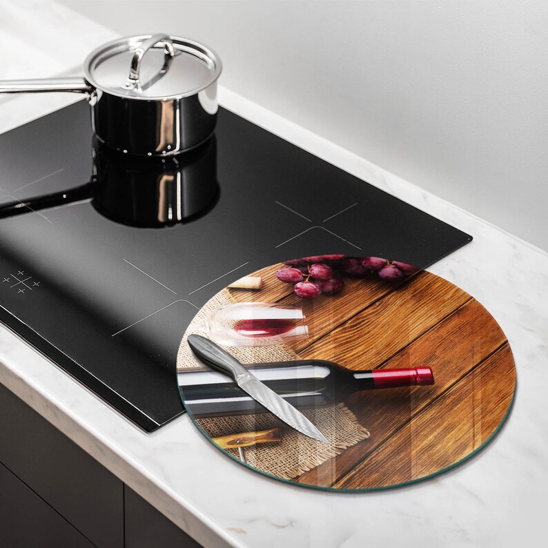 May include: A round glass cutting board with a wine theme. The board features a bottle of red wine, a glass of wine, grapes, and a knife on a wooden surface. A stainless steel pot sits on a black stovetop.
