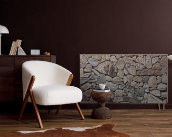 May include: A white armchair with wooden legs sits in a room with a dark brown wall. A stone-patterned wall panel is mounted on the wall. A small wooden stool with a white cup and saucer sits in front of the panel. A brown wooden dresser with a white lamp and books sits in the background.