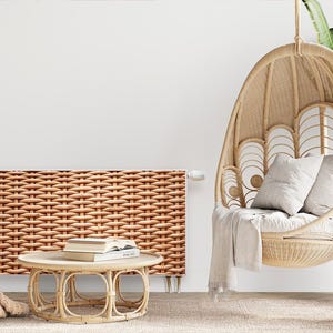 Op de afbeelding: Een witte hangstoel van gevlochten wilg met een beige kussen en een wit deken. De stoel hangt aan het plafond in een kamer met een witte muur en een radiatorbekleding van gevlochten wilg.