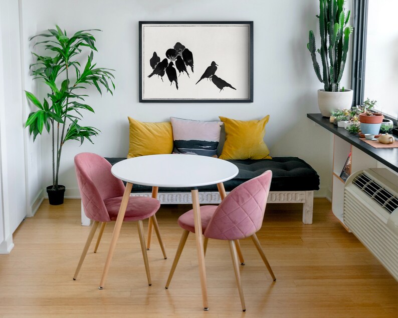 May include: A white round table with a wood base and two pink velvet chairs. The table is set in a living room with a black couch, yellow and pink throw pillows, and a black and white print of birds on the wall.