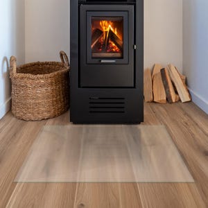 May include: A rectangular, transparent floor protector mat made of clear material, positioned on a light brown wood-effect floor in front of a black metal fireplace stove with a burning fire inside and a woven wicker basket nearby.