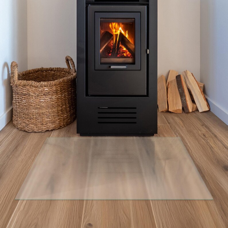 May include: A rectangular, transparent floor protector mat made of clear material, positioned on a light brown wood-effect floor in front of a black metal fireplace stove with a burning fire inside and a woven wicker basket nearby.