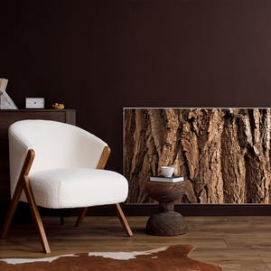 May include: A modern living room scene featuring a white armchair with wooden legs, a radiator cover with a tree bark design, and a small wooden side table. A white lamp and books sit on a dark brown cabinet. The room has a warm, inviting atmosphere.