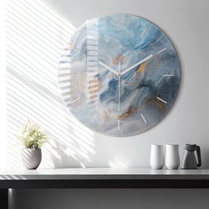 Può includere: Un orologio da parete circolare caratterizzato da un motivo astratto vorticoso nei toni del blu, grigio e oro. Il quadrante presenta una finitura liscia e lucida con sottili indicatori bianchi e semplici lancette bianche per l'ora.