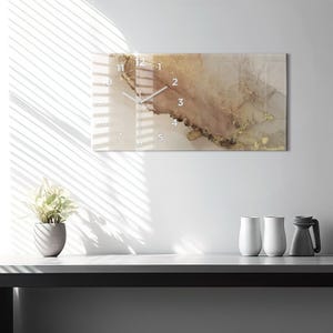 Peut inclure: Une horloge murale rectangulaire présentant un motif abstrait avec des tons tourbillonnants beige, brun et doré métallique. Le cadran affiche des chiffres blancs de 1 à 12 et deux aiguilles blanches sur une surface lisse et brillante marbrée.
