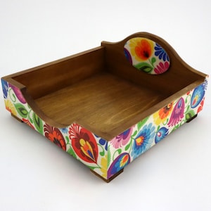 May include: A wooden napkin holder with a colourful floral design. The holder has a square base and a raised lip around the top.