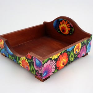 May include: A wooden napkin holder with a colourful floral design. The holder has a square base and a raised lip around the top. The floral design is in shades of red, orange, yellow, green, blue, and purple.