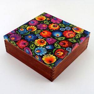 May include: A wooden box with a colourful floral pattern on the lid. The lid is made of a dark fabric with a black background and bright red, yellow, blue, and green flowers. The box is made of dark wood and has a natural finish.