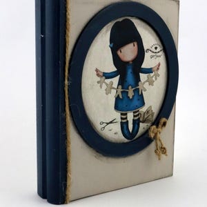 Puede incluir: Una caja de madera con un marco azul y un fondo blanco. La caja presenta un recorte circular con una ilustración colorida de una niña sosteniendo una cuerda de muñecas de papel. El texto "Gorjuss" es visible en la imagen.