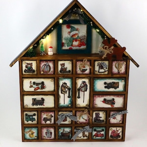 以下が含まれることがあります： 家型の木製クリスマスアドベントカレンダー。カレンダーには24個の小さな引き出しがあり、それぞれにヴィンテージのおもちゃのイラストが飾られています。上部には帽子をかぶったクマの小さな額入り写真があり、トナカイとライトが付いています。