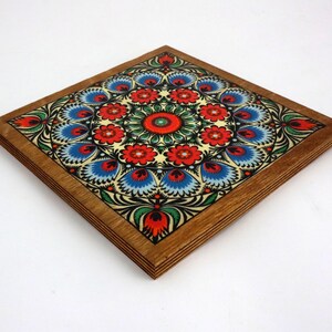 May include: A square wooden coaster featuring a colourful floral design. The design incorporates red, blue, green and yellow flowers on a cream background. The coaster is framed in a dark wood.