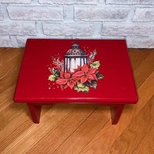 May include: A small, red wooden step stool with a decorative Christmas lantern and poinsettia design on the top. The stool has a rectangular top and angled legs. The background includes a brick wall and wooden floor.