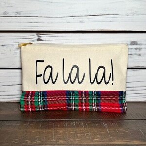 May include: Cream-colored zippered pouch with "Fa la la!" in black script. The bottom features a red, green, and blue plaid pattern. The pouch has a gold-colored zipper.