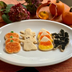 May include: A white oval platter holds an assortment of Halloween-themed sugar cookies. The selection includes orange pumpkin-shaped cookies, white ghost-shaped cookies, triangular candy corn-shaped cookies, and black cat-shaped cookies.