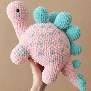 Könnte beinhalten: Ein rosa und blau gehäkeltes Dinosaurier-Spielzeug. Das Plüschtier hat einen rosa Körper mit blauen Akzenten, einschließlich Flecken und Rückenplatten. Der Dinosaurier wird vor einem neutralen Hintergrund gehalten.