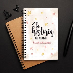 May include: Spiral-bound notebook with the text "La historia de mi vida" in black script, accented with gold and pink stars. A brown notebook and a black pen are also visible.