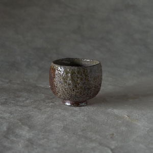 May include: A handmade ceramic bowl with a textured surface. The bowl is a mix of brown and green hues, with a small, rounded base. The interior is dark, and the bowl has a rustic, organic aesthetic.