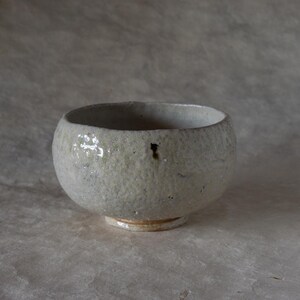 May include: A handcrafted, off-white ceramic bowl with a rounded form and a small, circular base. The bowl has a textured surface and a small hole on the side. The bowl is likely used for serving food or as a decorative item.