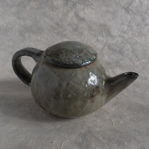 Può includere: Una piccola teiera rotonda in ceramica verde e nera con un coperchio. La teiera ha un beccuccio e un manico.