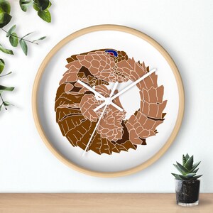 May include: A round wall clock with a light wood frame and a white face. The center features a stylized graphic illustration of a brown pangolin curled into a circular shape. The clock has three simple white hands indicating the time.