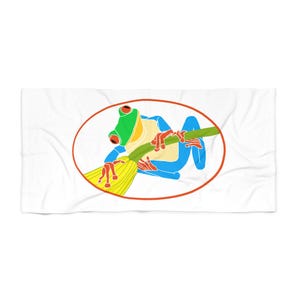 May include: A white rectangular beach towel featuring a centered orange oval frame containing a graphic illustration of a red-eyed tree frog with a green head, blue limbs, and a yellow body, perched on a green branch with yellow leaves.