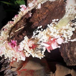 May include: A delicate floral crown with white and pink dried flowers. The crown is made with small white flowers and pink flowers with a few sprigs of dried grass.