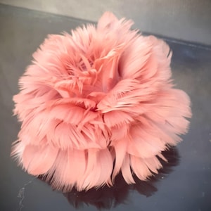 May include: A fluffy, round, peach-colored feather flower. The feathers are layered, creating a full, textured appearance. The flower is centered in the image, with a dark, reflective surface beneath it.