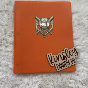 May include: A bright orange binder featuring a central graphic of a baseball with the name Collin printed in white, positioned over two crossed baseball bats. A wooden cutout sign with the text Kinsley DOWDY CO is placed on the lower right.