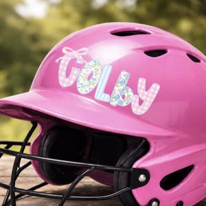 May include: A bright pink softball helmet with a black faceguard. The name "Colby" is displayed on the side in a decorative font with floral and gingham patterns. The helmet sits on a wooden surface.