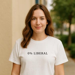 May include: A white, short-sleeved t-shirt with the text "0% LIBERAL" printed in black. The shirt is worn by a person with brown hair, smiling, and standing outdoors.