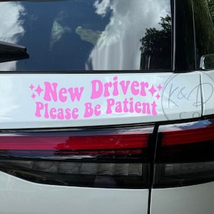 May include: A pink decal on the back of a white car. The decal reads "New Driver Please Be Patient" in a retro font with starburst accents. The car's taillight is visible.