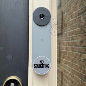 May include: A gray doorbell camera with a black lens and a white sticker that says "NO SOLICITING" on a white door frame.