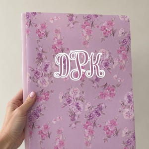 May include: A rectangular notebook with a light purple cover featuring a repeating floral pattern in shades of pink and violet. The center of the cover displays the white stylized monogram letters DPK in a decorative, cursive-style font.