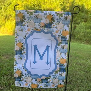 K&ouml;nnte beinhalten: Eine rechteckige Gartenflagge mit einem sich wiederholenden Muster aus orangefarbenen und hellblauen K&uuml;rbissen, wei&szlig;en Blumen und blauen Karomustern. In der Mitte umgibt ein hellblauer, gewellter Rahmen einen gro&szlig;en, blauen Buchstaben M.