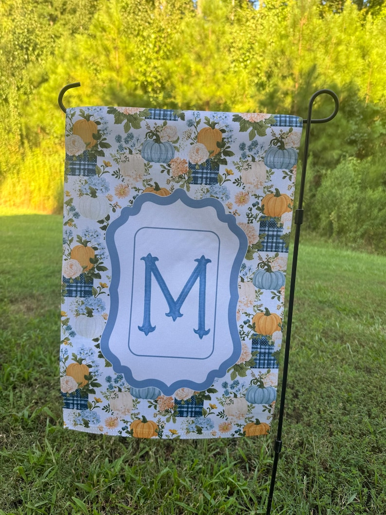 K&ouml;nnte beinhalten: Eine rechteckige Gartenflagge mit einem sich wiederholenden Muster aus orangefarbenen und hellblauen K&uuml;rbissen, wei&szlig;en Blumen und blauen Karomustern. In der Mitte umgibt ein hellblauer, gewellter Rahmen einen gro&szlig;en, blauen Buchstaben M.