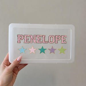 May include: A rectangular, translucent white plastic storage box lid featuring the name PENELOPE in pink varsity-style letters with a dark outline. Below the name are five glittery stars in light blue, light pink, teal, purple, and lime green.