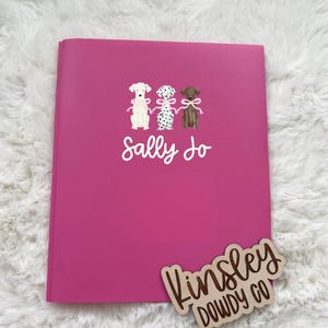 May include: A bright pink notebook with the name "Sally Jo" in white script. The notebook features illustrations of three dogs with bows. A wooden sign that says "Kinsley Dowdy Co" is in the bottom right corner.
