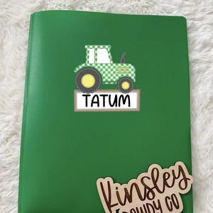 May include: Green notebook with a tractor graphic. The tractor is green and white checkered with yellow wheels. The name "TATUM" is printed on a white label. A wooden cutout with the name "Kinsley DOWDY CO" is also included.